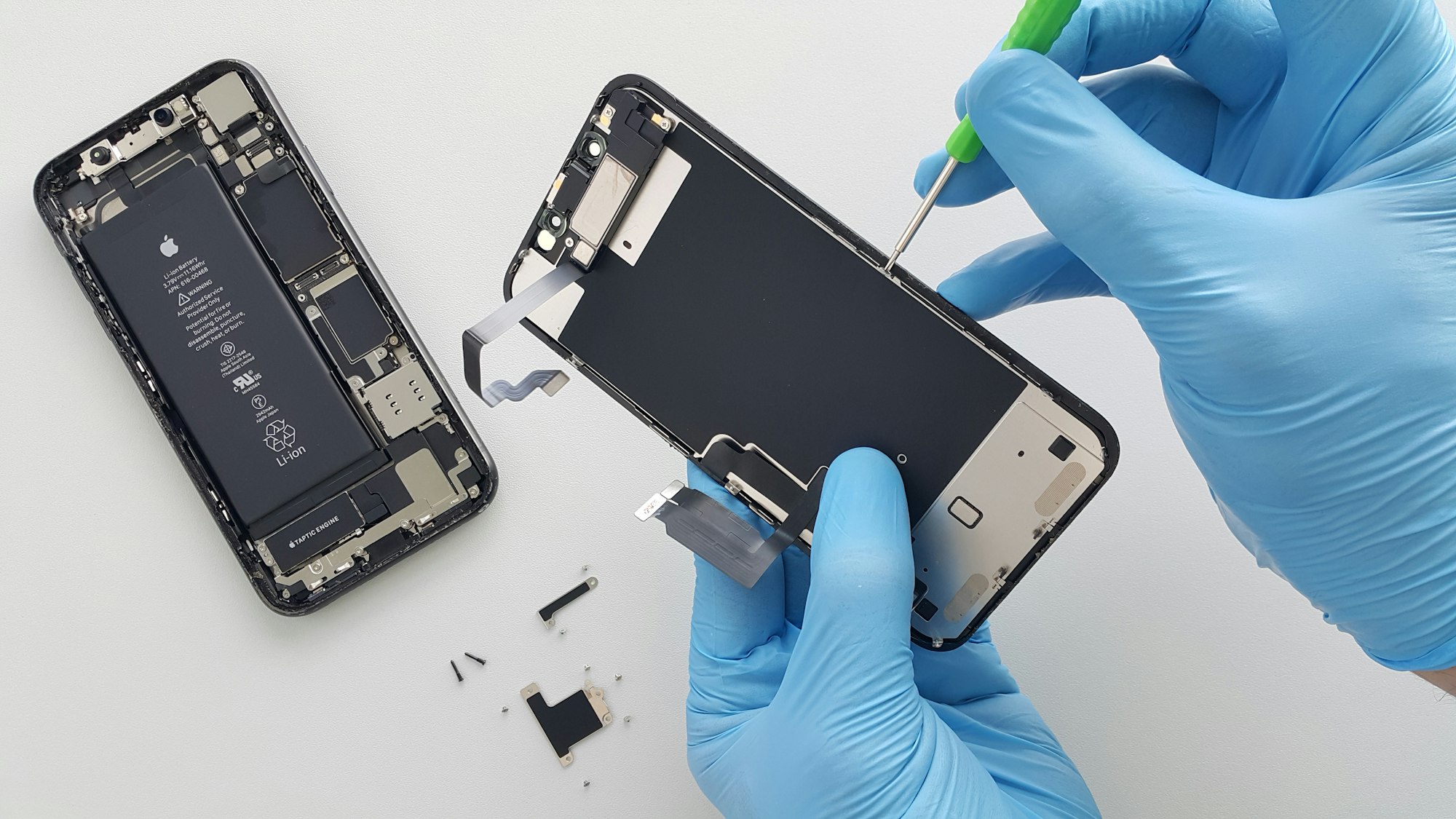 Technicien ganté soulevant l'écran d'un iPhone sur un plan de travail blanc.